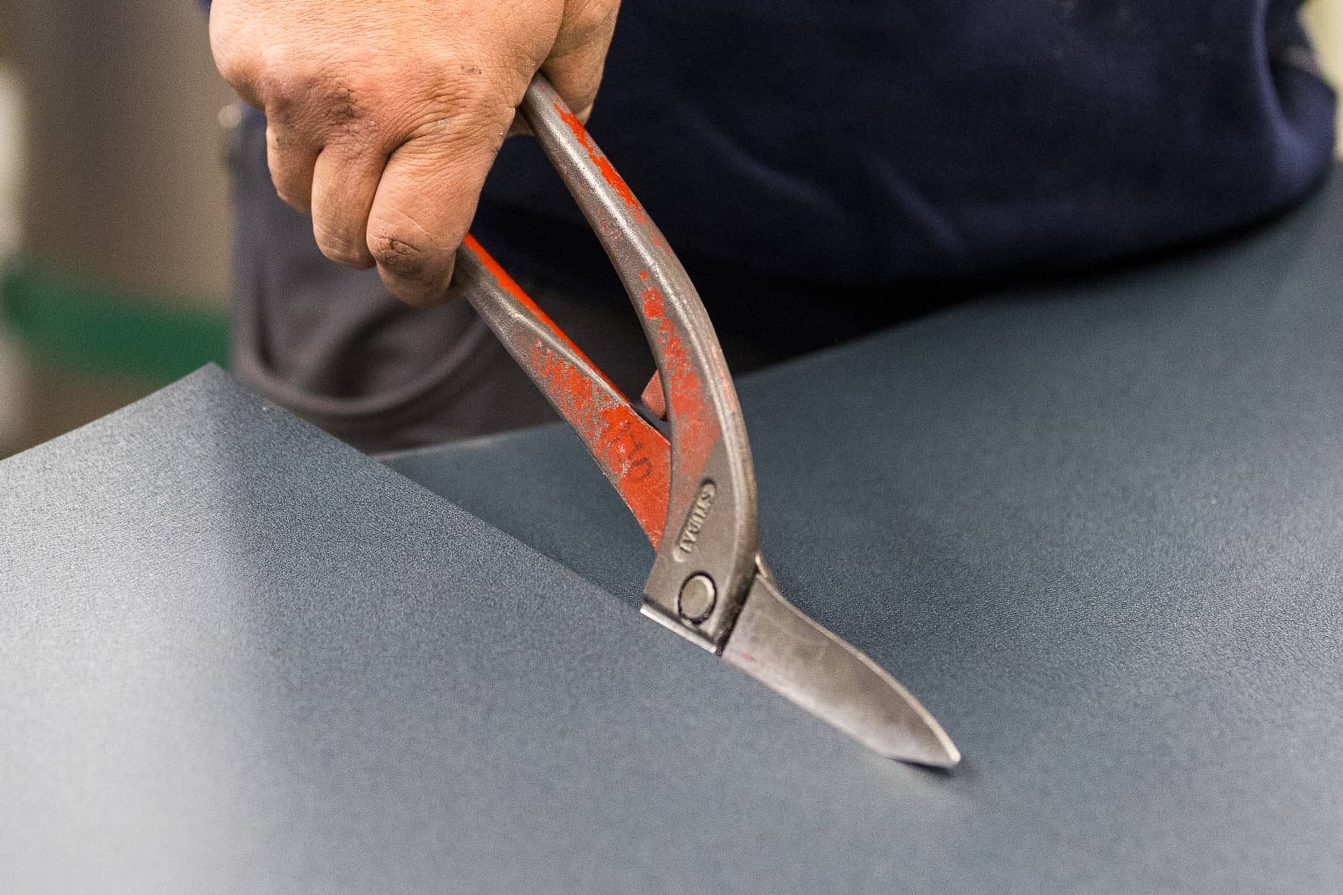 Das Bild zeigt einen Ausschnitt mit einer Hand an einer Metallschere beim Schneiden von einer Metallplatte.